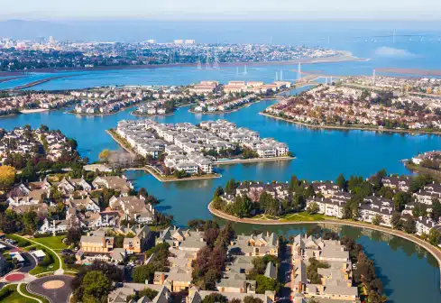Aerial view of a coastal residential area with houses surrounding blue waterways. The landscape features numerous channels and peninsulas, with distant views of a city and mountains under a clear sky.