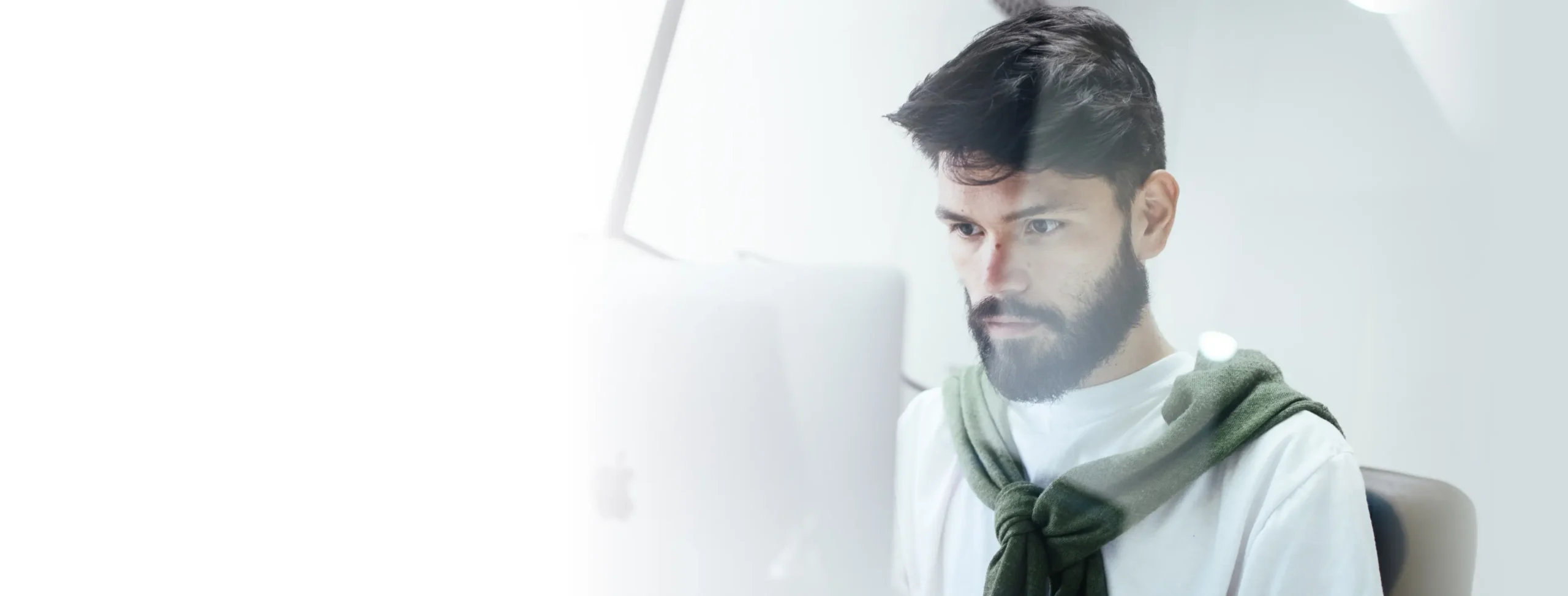 A man with a beard and a green sweater draped over his shoulders is focused on a computer screen. The image is partially overexposed, creating a soft white fade on the left side.