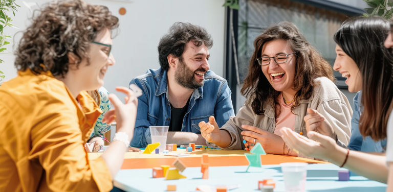 A group of five people sit around a colorful table playing a board game. They are smiling and laughing, enjoying the sunny outdoor setting. The table is scattered with game pieces and drinks.