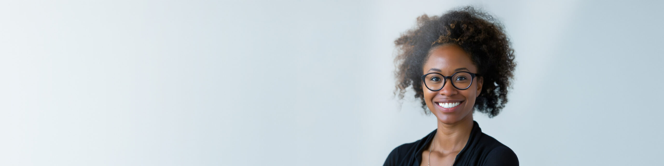 Smiling person with curly hair and glasses stands against a plain light background. They are wearing a black top and a cardigan. The image has ample blank space on the left side.