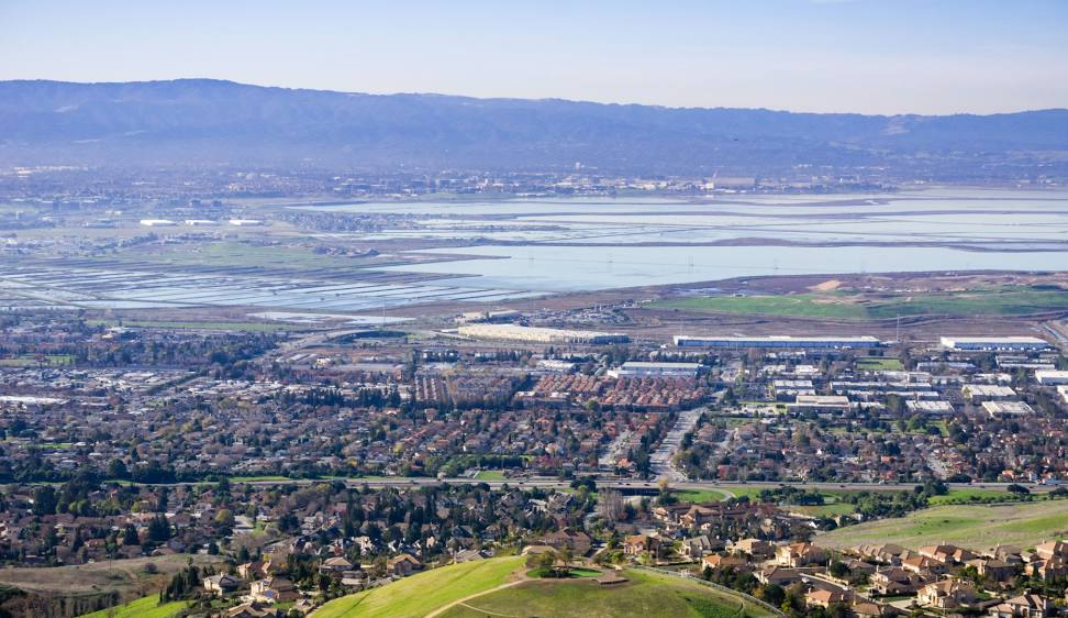 Aerial view of a suburban neighborhood with houses, buildings, and roads, set against a backdrop of wetlands and distant hills under a clear sky.